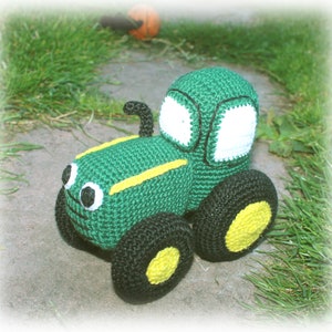 May include: A green crocheted toy tractor with black wheels and yellow accents. The tractor has a white window and black eyes.