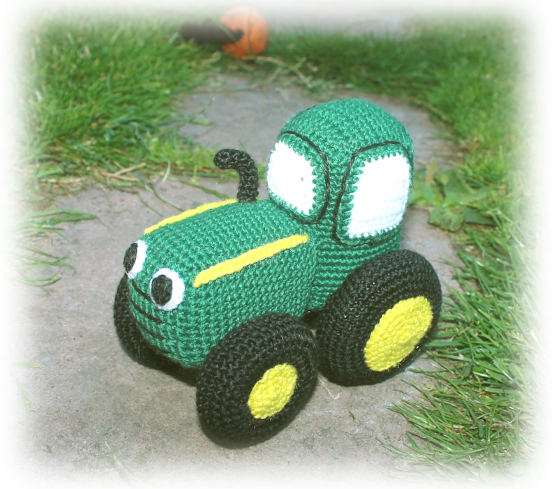 May include: A green crocheted toy tractor with black wheels and yellow accents. The tractor has a white window and black eyes.