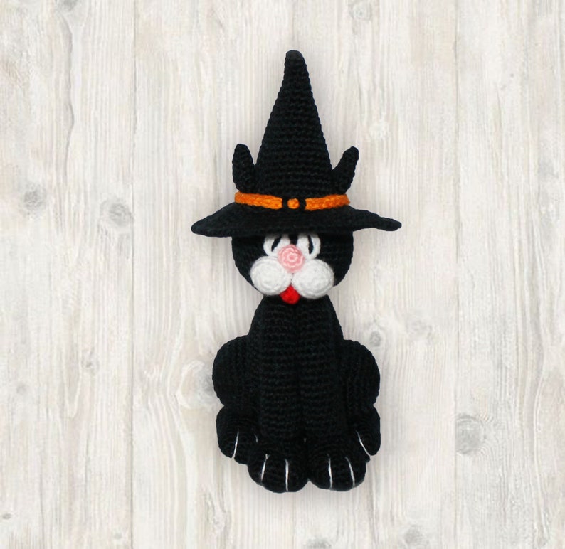 May include: A black crocheted cat wearing a witch's hat with an orange band. The cat has white facial features and a pink nose.