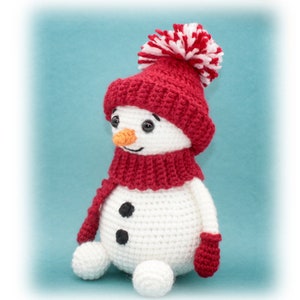 Könnte beinhalten: Ein weißer gehäkelter Schneemann, der eine rote Strickmütze mit einem weißen Bommel und einen roten Schal trägt. Der Schneemann hat schwarze Augen, eine Karottennase und zwei schwarze Knöpfe.