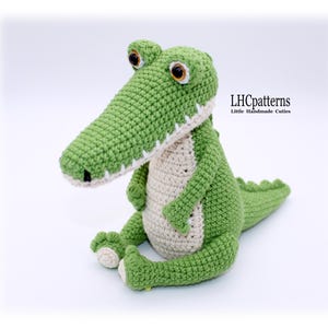 Pode incluir: Um brinquedo de crochê verde e creme feito à mão, em forma de jacaré, com olhos grandes e expressivos e um focinho longo. O jacaré tem uma barriga creme e dentes brancos. O texto "LHCpatterns Little Handmade Cuties" é visível.