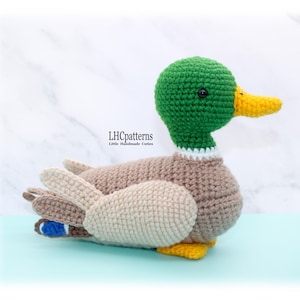 Könnte beinhalten: Eine handgefertigte Häkelente mit grünem Kopf, gelbem Schnabel und braunem Körper. Die Ente hat weiße Akzente um den Hals und an den Flügeln. Die Flügel sind beige und die Schwanzfedern blau. Der Text "LHCpatterns" ist sichtbar.