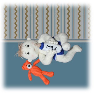 Op de afbeelding: Een gehaakte babypop met blond haar ligt op haar rug en houdt een witte fles vast met het woord "MILK" erop gedrukt. Een kleine oranje gehaakte teddybeer ligt naast de pop.