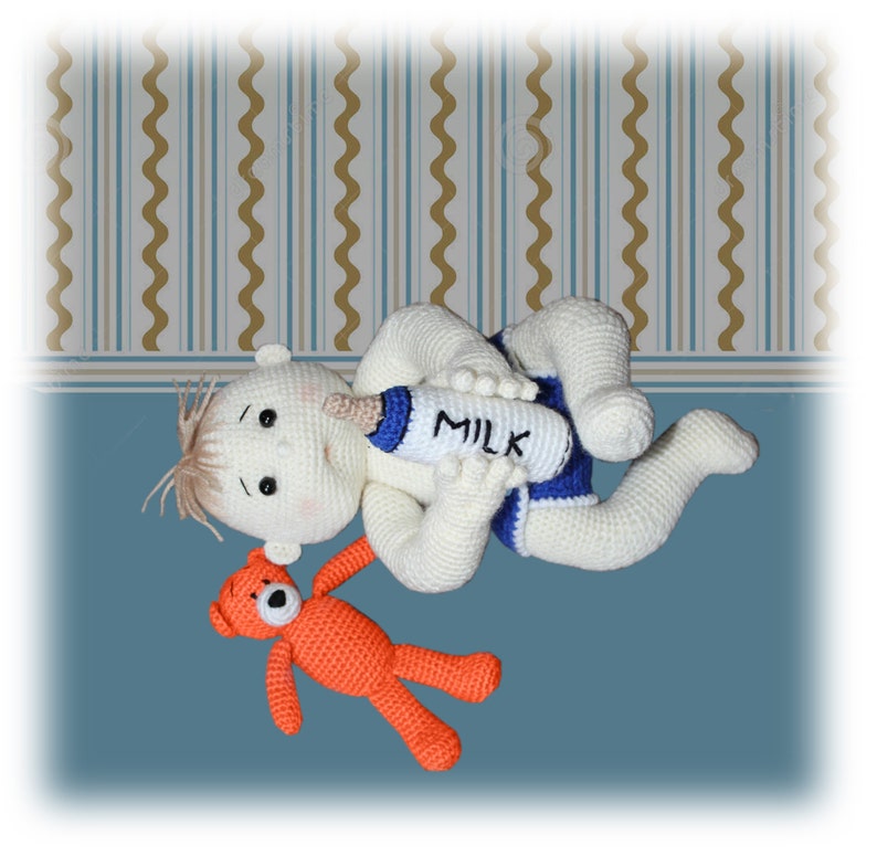 Op de afbeelding: Een gehaakte babypop met blond haar ligt op haar rug en houdt een witte fles vast met het woord "MILK" erop gedrukt. Een kleine oranje gehaakte teddybeer ligt naast de pop.