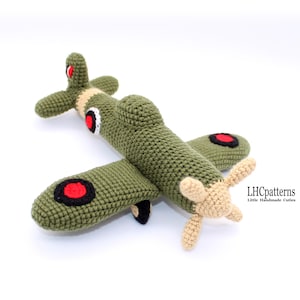 Puede incluir: Juguete de avión de crochet verde con círculos rojos y negros en las alas y una hélice de color beige. El juguete está hecho por LHCpatterns, Little Handmade Cuties.