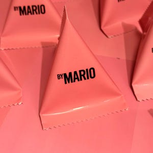 May include: Pink triangular packages arranged on a pink surface. Each package has the text "by MARIO" printed in black. The packages are folded into a pyramid shape.