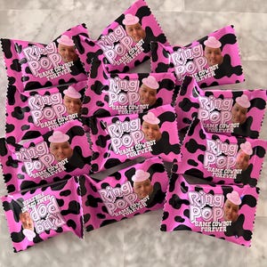 May include: A collection of Ring Pop candy wrappers featuring a pink and black cow print pattern. Each wrapper displays the text Ring Pop and Same Cowboy Forever, along with a photographic portrait of a person wearing a pink cowboy hat.