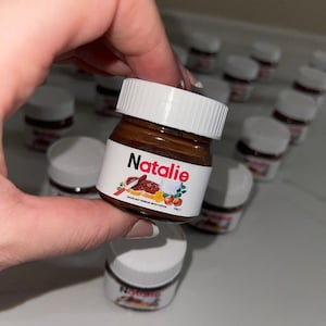 Op de afbeelding: Een klein potje hazelnootpasta met de naam "Natalie" op het etiket. Het potje is bruin met een wit deksel en is omringd door andere potjes.