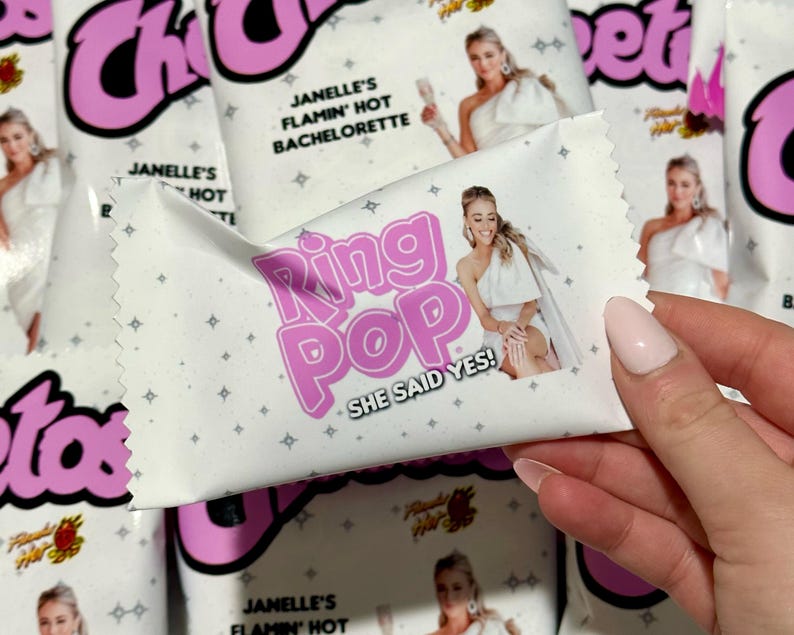 May include: A white rectangular candy wrapper with pink text that reads Ring Pop and She Said Yes. The wrapper features a photograph of a person in a white one-shoulder dress and a pattern of small gray stars. The text Janelle's Flamin' Hot Bachelorette is printed above the image.