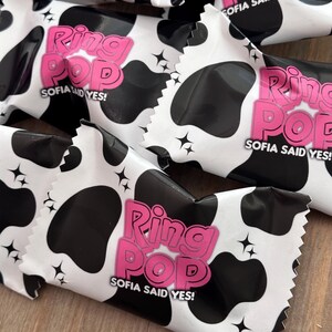 May include: Multiple Ring Pop candy wrappers featuring a black and white cow print pattern. Each wrapper displays the pink text Ring Pop and the white text Sofia Said Yes, accented with small black four-pointed star icons across the surface.