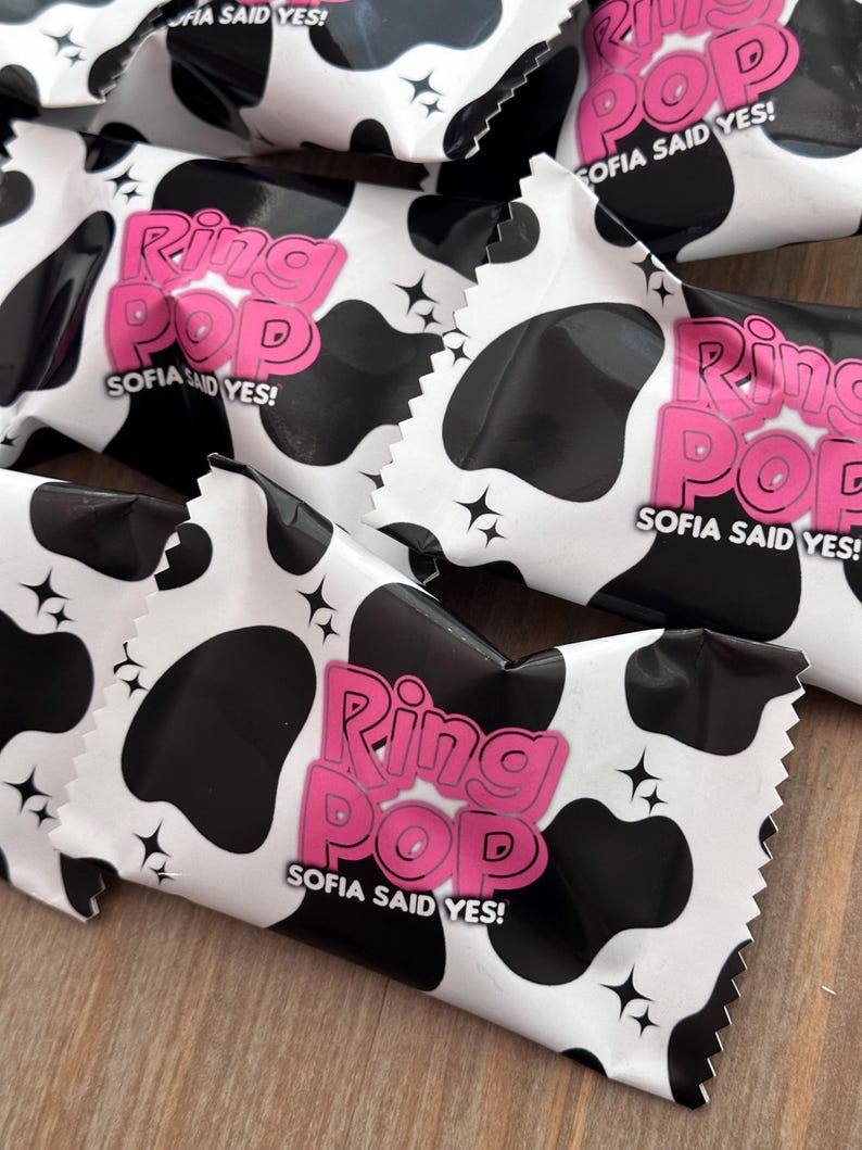 May include: Multiple Ring Pop candy wrappers featuring a black and white cow print pattern. Each wrapper displays the pink text Ring Pop and the white text Sofia Said Yes, accented with small black four-pointed star icons across the surface.