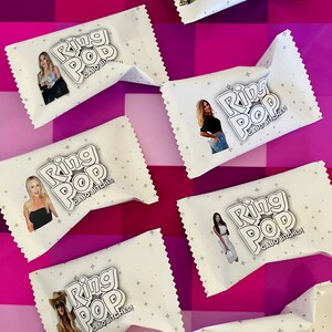 May include: A collection of white rectangular candy wrappers featuring the text Ring Pop and Cabo Bitches! alongside various photographic portraits of individuals. Each wrapper is decorated with a subtle pattern of small grey stars.