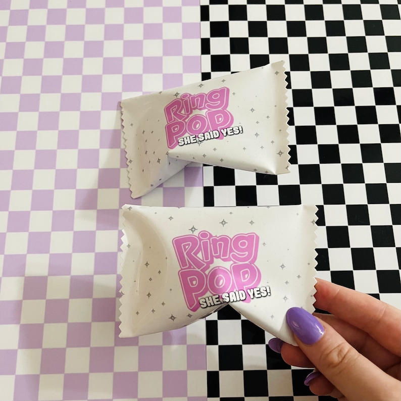 May include: Two white rectangular candy wrappers with serrated edges featuring the pink text Ring Pop and the black text SHE SAID YES! accented by small grey star icons. The wrappers are made of a smooth material and have a crimped seal design.