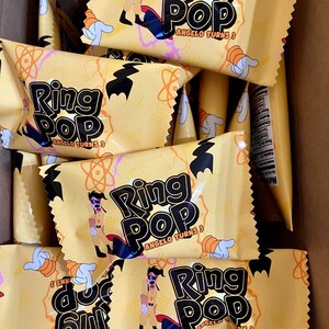 May include: A collection of yellow Ring Pop candy wrappers featuring a cartoon character in a tan outfit, black gloves, and a red cape. The text reads Ring Pop and Angelo Turns 3, accompanied by orange atom-like graphics and white gloved hand icons.