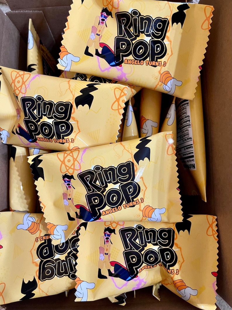 May include: A collection of yellow Ring Pop candy wrappers featuring a cartoon character in a tan outfit, black gloves, and a red cape. The text reads Ring Pop and Angelo Turns 3, accompanied by orange atom-like graphics and white gloved hand icons.