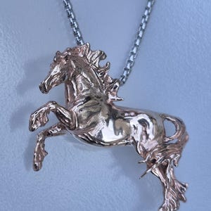 Könnte beinhalten: Eine roségoldfarbene Anhängerkette mit einem steigenden Pferd. Das Pferd ist sehr detailliert, mit fließender Mähne und Schweif. Der Anhänger hängt an einer silberfarbenen Kette. Die Kette ist in Zentimetern bemessen.
