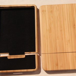 May include: A bamboo phone case with a black felt interior. The case has a hinged lid and a slot for holding a phone.