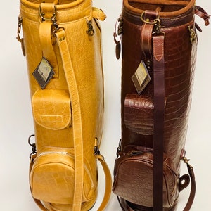 May include: Two golf bags in the image. One is a light tan color, and the other is a dark brown. Both bags have a crocodile skin texture, multiple pockets, and shoulder straps. The bags have metal hardware and tags.