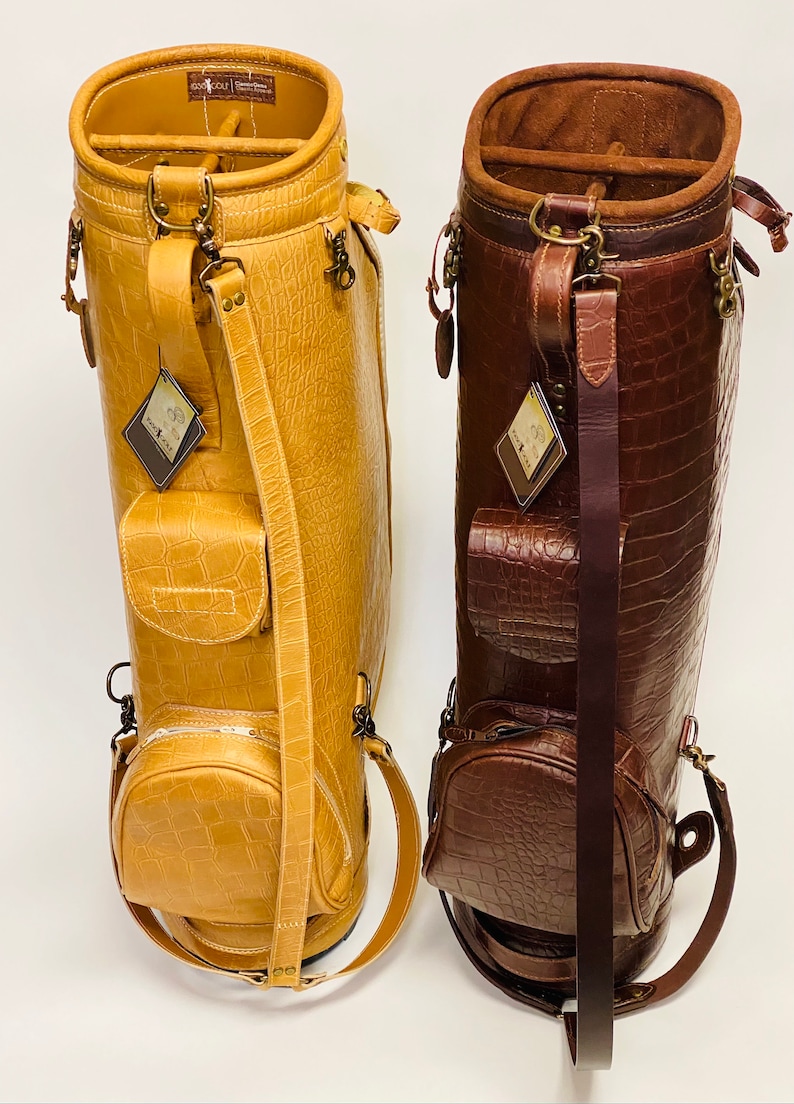 May include: Two golf bags in the image. One is a light tan color, and the other is a dark brown. Both bags have a crocodile skin texture, multiple pockets, and shoulder straps. The bags have metal hardware and tags.