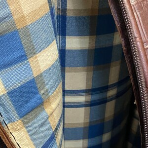 May include: Close-up of a brown leather bag with a plaid interior. The interior fabric features a blue, beige, and white plaid pattern. The bag has a zipper closure and a textured exterior.