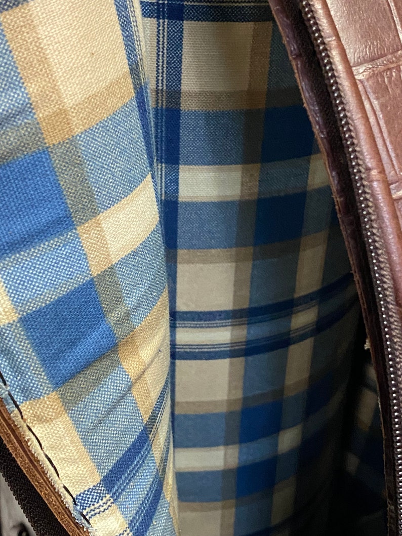 May include: Close-up of a brown leather bag with a plaid interior. The interior fabric features a blue, beige, and white plaid pattern. The bag has a zipper closure and a textured exterior.