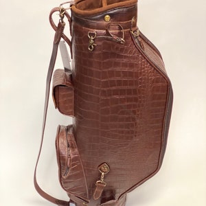 May include: A brown golf bag with a crocodile skin texture. The bag has multiple pockets, a shoulder strap, and metal hardware. The interior is lined with a lighter brown suede-like material.