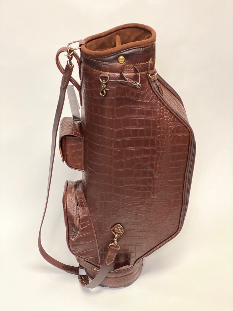 May include: A brown golf bag with a crocodile skin texture. The bag has multiple pockets, a shoulder strap, and metal hardware. The interior is lined with a lighter brown suede-like material.