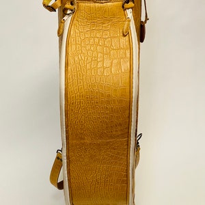 May include: A tan golf bag with a textured, crocodile-like pattern. The bag has white accents, a zipper, and a handle. The bag is made of leather and has a sturdy base. The bag is designed to carry golf clubs.