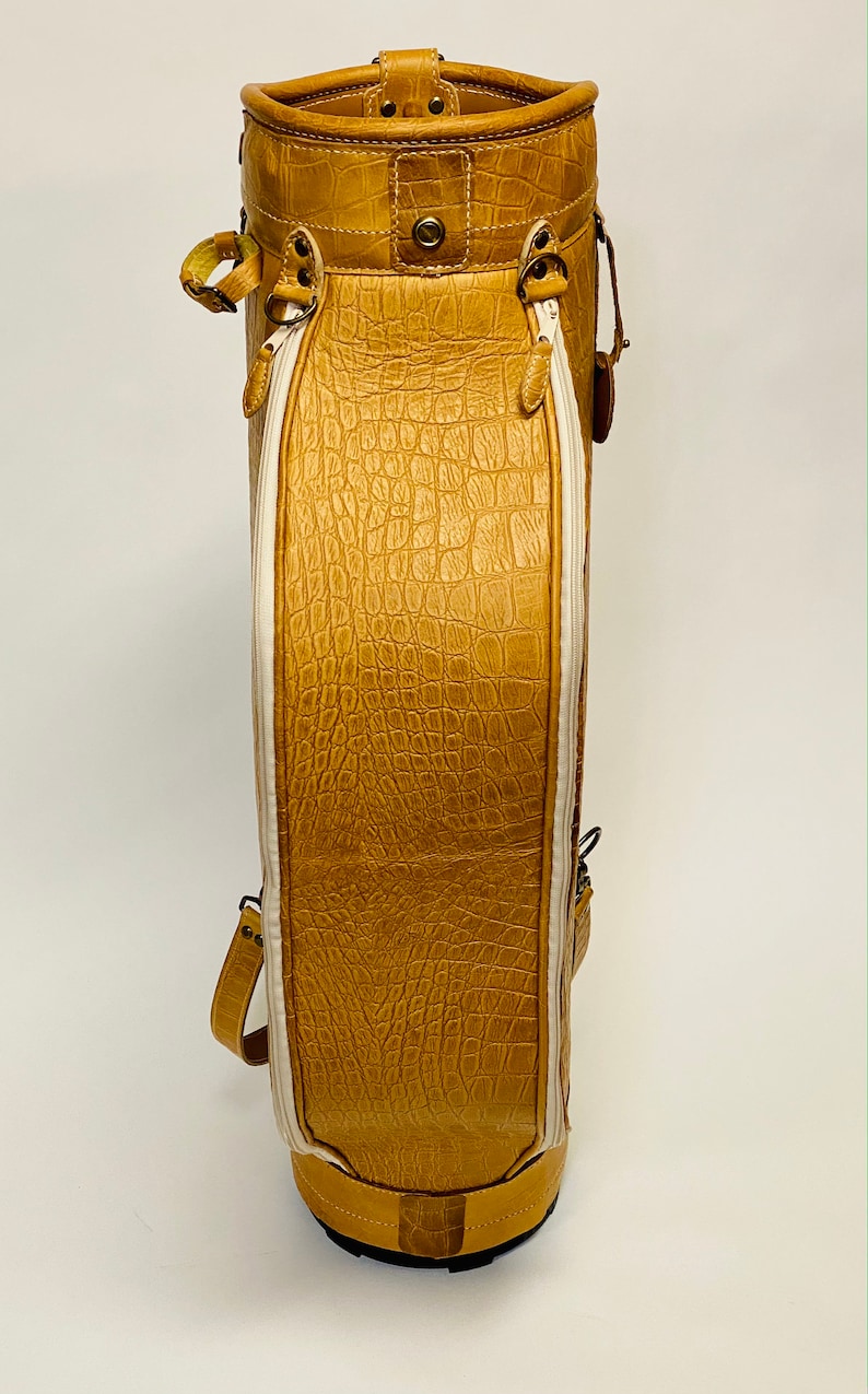 May include: A tan golf bag with a textured, crocodile-like pattern. The bag has white accents, a zipper, and a handle. The bag is made of leather and has a sturdy base. The bag is designed to carry golf clubs.