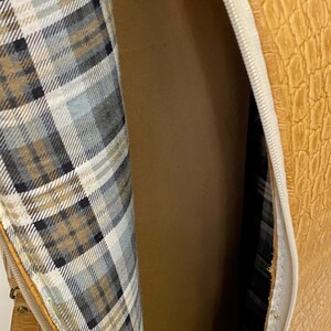 May include: Open view of a tan, textured bag with a zipper. The interior lining features a plaid pattern in shades of gray, white, and tan. The bag's exterior has a reptile-like texture.