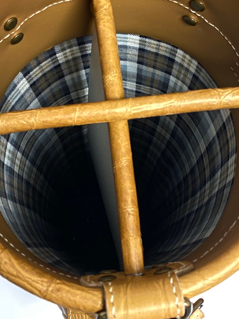 May include: Tan leather golf bag with a plaid interior. The bag has a cross-shaped divider at the top and is stitched with white thread. The interior is a plaid pattern of black, white, and brown.