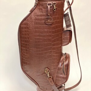May include: A brown leather golf bag with a crocodile skin texture. The bag has multiple pockets, a shoulder strap, and metal hardware. The top of the bag has a brown suede lining and the brand name "1936 Golf" is visible.
