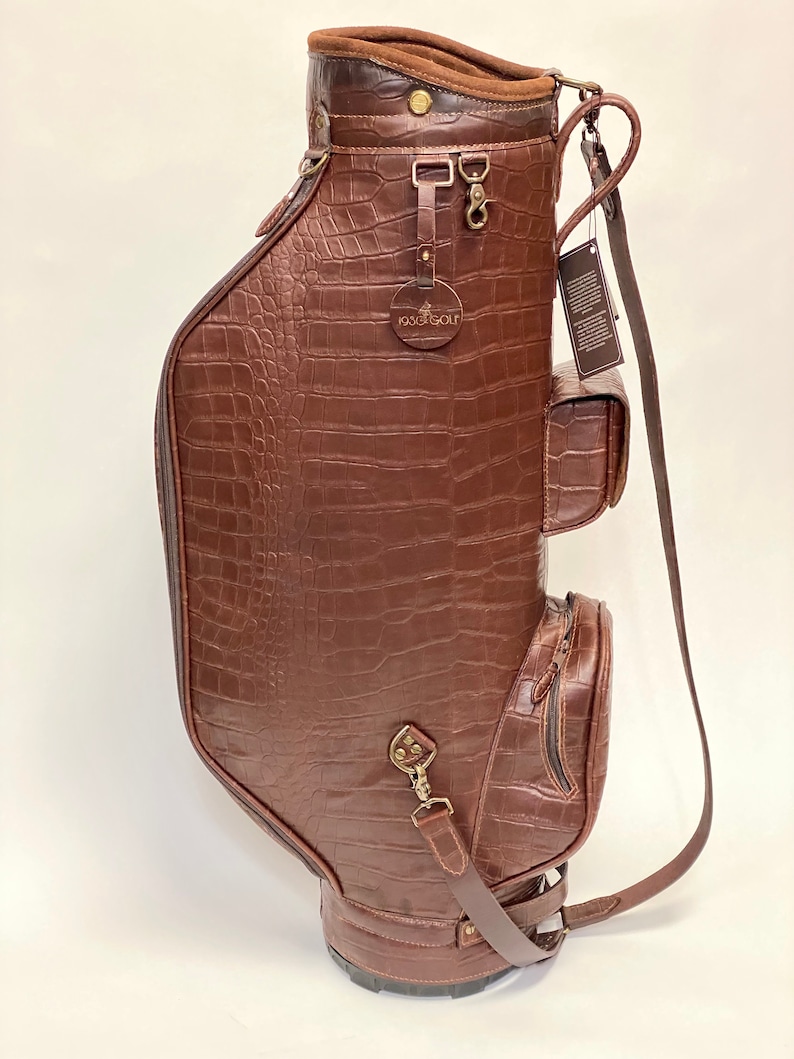 May include: A brown leather golf bag with a crocodile skin texture. The bag has multiple pockets, a shoulder strap, and metal hardware. The top of the bag has a brown suede lining and the brand name "1936 Golf" is visible.