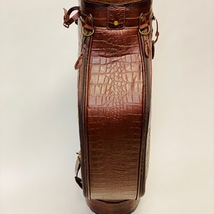 May include: A brown leather golf bag with a crocodile skin texture. The bag has a cylindrical shape with a rounded base and a suede-lined top. It features brass hardware, zippers, and straps.