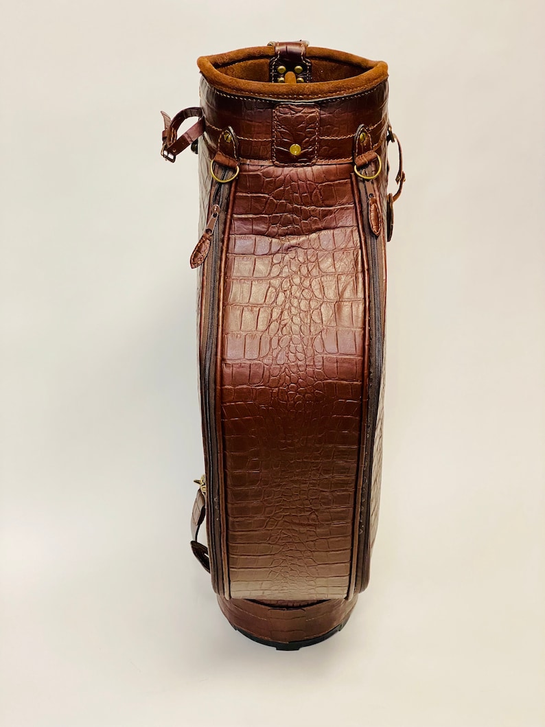 May include: A brown leather golf bag with a crocodile skin texture. The bag has a cylindrical shape with a rounded base and a suede-lined top. It features brass hardware, zippers, and straps.