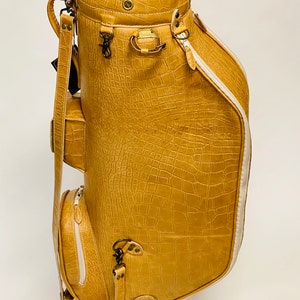 May include: Tan golf bag with a crocodile skin texture. The bag has multiple pockets, a white zipper, and a white stripe down the side. It features metal hardware and a shoulder strap. The bag is designed to carry golf clubs.