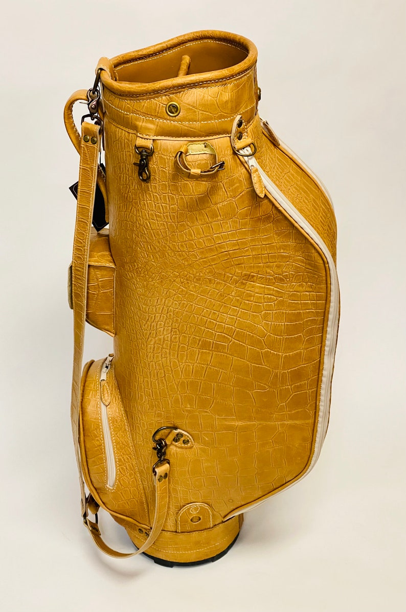 May include: Tan golf bag with a crocodile skin texture. The bag has multiple pockets, a white zipper, and a white stripe down the side. It features metal hardware and a shoulder strap. The bag is designed to carry golf clubs.