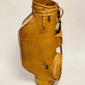 May include: Tan golf bag with a crocodile skin texture. The bag has multiple pockets, a shoulder strap, and a white zipper. The bag has the text "Jaxx Golf" on the front. The bag is on a white background.