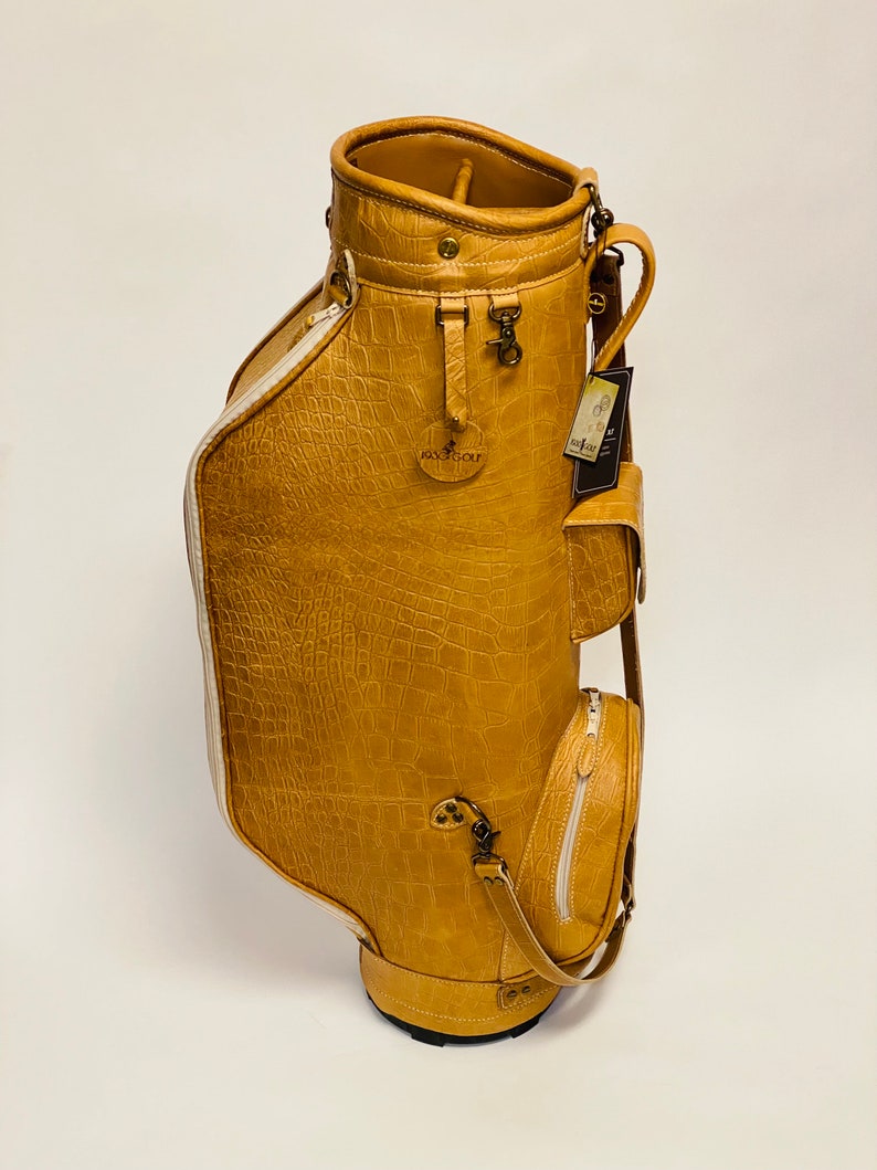 May include: Tan golf bag with a crocodile skin texture. The bag has multiple pockets, a shoulder strap, and a white zipper. The bag has the text "Jaxx Golf" on the front. The bag is on a white background.