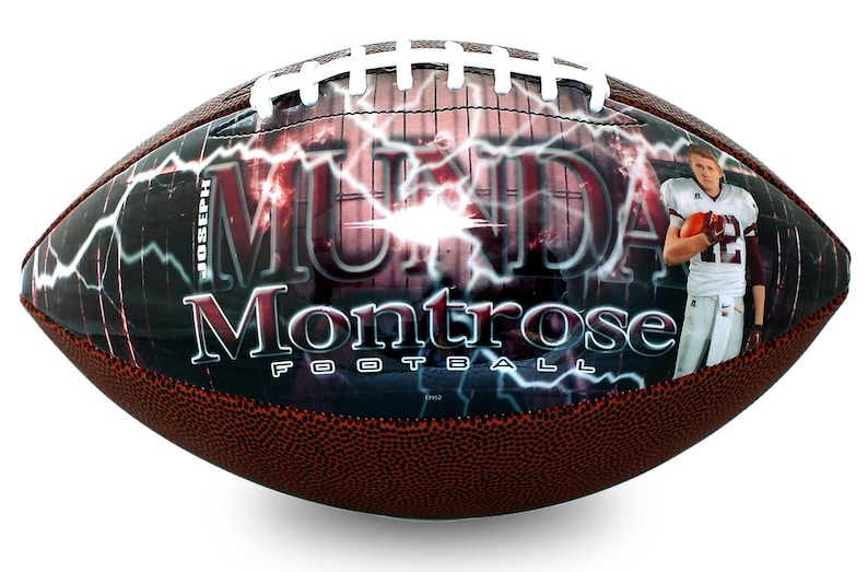 Op de afbeelding: Een bruine en witte voetbal met een zwart en rood ontwerp. Het ontwerp bevat de naam van een speler, "MUNDAY", en de tekst "Montrose Football". Het ontwerp bevat ook een bliksemontwerp en een foto van een speler.