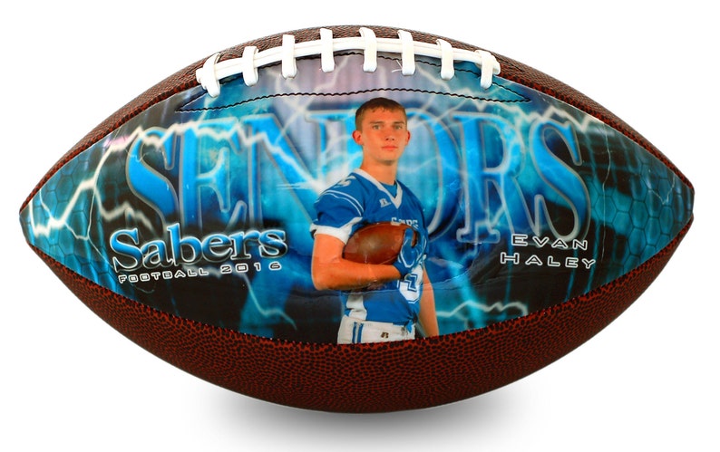 Op de afbeelding: Een bruine en witte voetbal met een blauw en wit ontwerp. Het ontwerp heeft een bliksem patroon, het woord "Seniors" in blauw en de woorden "Sabers Football 2016". Een foto van een jonge man in een blauw voetbaluniform bevindt zich in het midden van de voetbal.