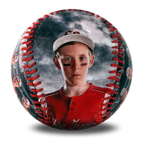 May include: A baseball with a photo of a young boy wearing a red jersey and a baseball cap. The baseball has a grey and white background with a cloudy sky. The boy is looking at the camera with a serious expression. The baseball has red stitching and the text "King Trevor" is visible at the bottom.
