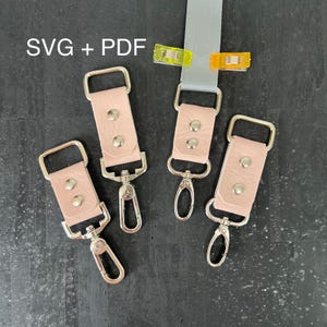 May include: Four pink leather keychains with silver swivel clips. Each keychain has two silver rivets. The keychains are arranged on a black surface. SVG + PDF text is in the top left corner of the image.