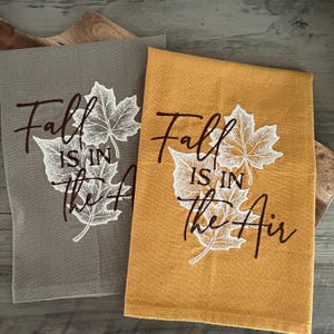 May include: Two rectangular kitchen towels, one in gray and one in mustard yellow, each featuring white embroidered maple leaf designs and the dark brown text "Fall IS IN The Air" printed in a combination of script and block lettering.