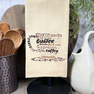 May include: A beige kitchen towel with brown embroidery of a coffee cup and the words "Espresso", "latte", "mocha", "coffee", "Mocha French Roast", "Espresso", "latte", "coffee", "Cafe Mocha", "Cappuccino", and "COFFEE".