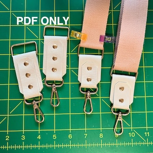 May include: Three white leather straps with gold metal hardware and clasps. The straps have three evenly spaced gold dots on each strap. The straps are on a green cutting mat with a ruler marked in inches.