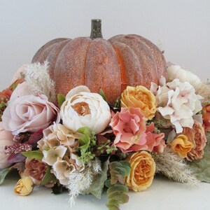 K&ouml;nnte beinhalten: Ein Herbst-Blumenarrangement mit einer k&uuml;nstlichen K&uuml;rbis, verschiedenen Herbstfarben wie Pfirsich-, Creme- und Orangenrosen sowie wei&szlig;en Hortensien. Der K&uuml;rbis ist von Gr&uuml;npflanzen und getrockneten Gr&auml;sern umgeben.