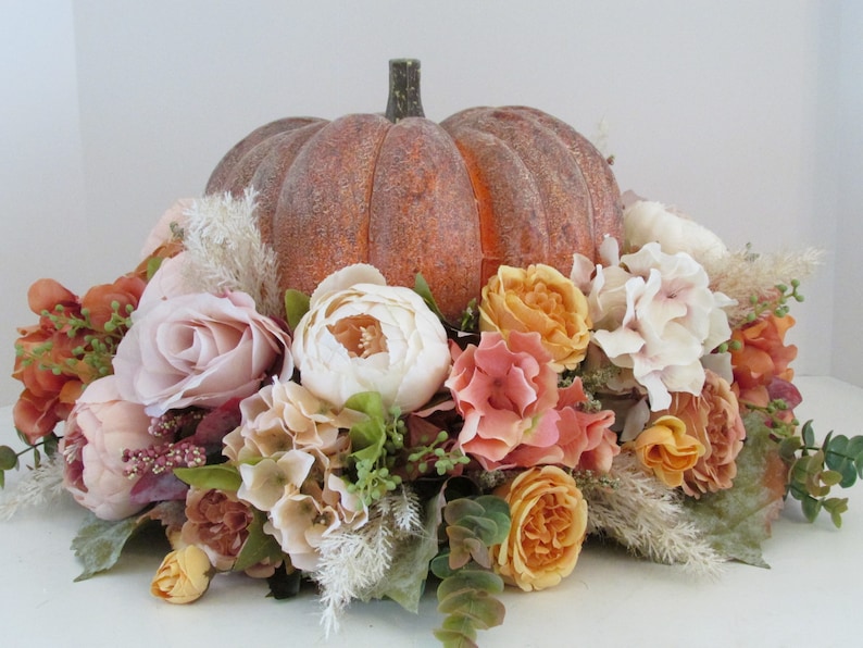 K&ouml;nnte beinhalten: Ein Herbst-Blumenarrangement mit einer k&uuml;nstlichen K&uuml;rbis, verschiedenen Herbstfarben wie Pfirsich-, Creme- und Orangenrosen sowie wei&szlig;en Hortensien. Der K&uuml;rbis ist von Gr&uuml;npflanzen und getrockneten Gr&auml;sern umgeben.
