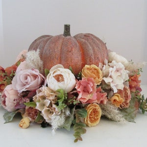 K&ouml;nnte beinhalten: Ein k&uuml;nstlicher K&uuml;rbis-Mittelpunkt, der mit einer Vielzahl von Herbstblumen geschm&uuml;ckt ist, darunter Rosen, Pfingstrosen und Hortensien. Der K&uuml;rbis ist von Gr&uuml;n und getrockneten Gr&auml;sern umgeben.