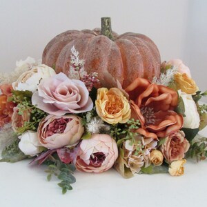K&ouml;nnte beinhalten: Ein k&uuml;nstlicher K&uuml;rbis-Mittelpunkt mit einer Vielzahl von Herbstblumen in Pfirsich-, Rosa- und Braunt&ouml;nen. Der K&uuml;rbis ist von einem Ring aus Blumen und Gr&uuml;n umgeben.